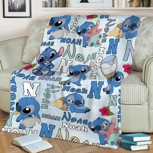 Może przedstawiać: Jasnoniebieski koc z postacią z kresk&oacute;wki Stitch w r&oacute;żnych pozach. Na kocu nadrukowano imię "Noah" i literę "N". Czerwone kwiaty i butelka dla dziecka są r&oacute;wnież widoczne.