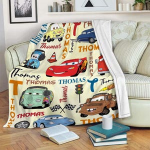 May include: A cream-colored throw blanket featuring cartoon cars and the name "Thomas" in various fonts. The blanket showcases a red race car, a blue car, and a brown tow truck, along with other vehicles and racing-themed elements.