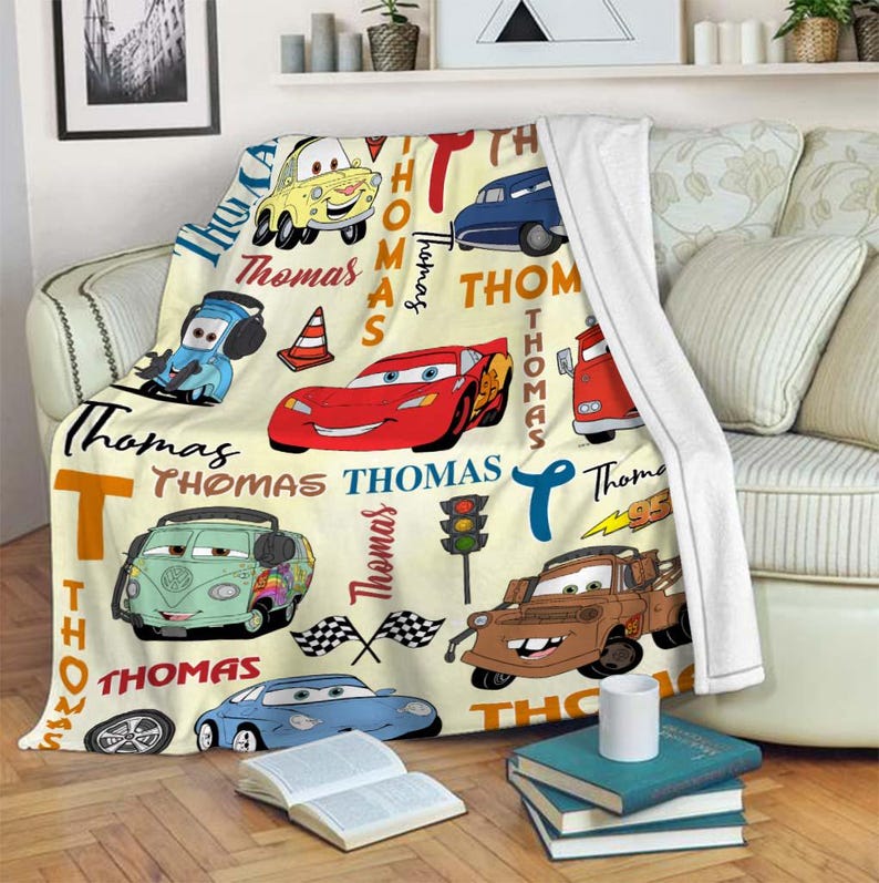 May include: A cream-colored throw blanket featuring cartoon cars and the name "Thomas" in various fonts. The blanket showcases a red race car, a blue car, and a brown tow truck, along with other vehicles and racing-themed elements.