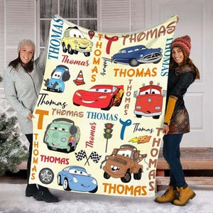 May include: A cream-colored blanket featuring cartoon cars and the name "Thomas" in various fonts and colors. The blanket showcases images of a yellow car, a blue car, a red race car, and a brown tow truck.