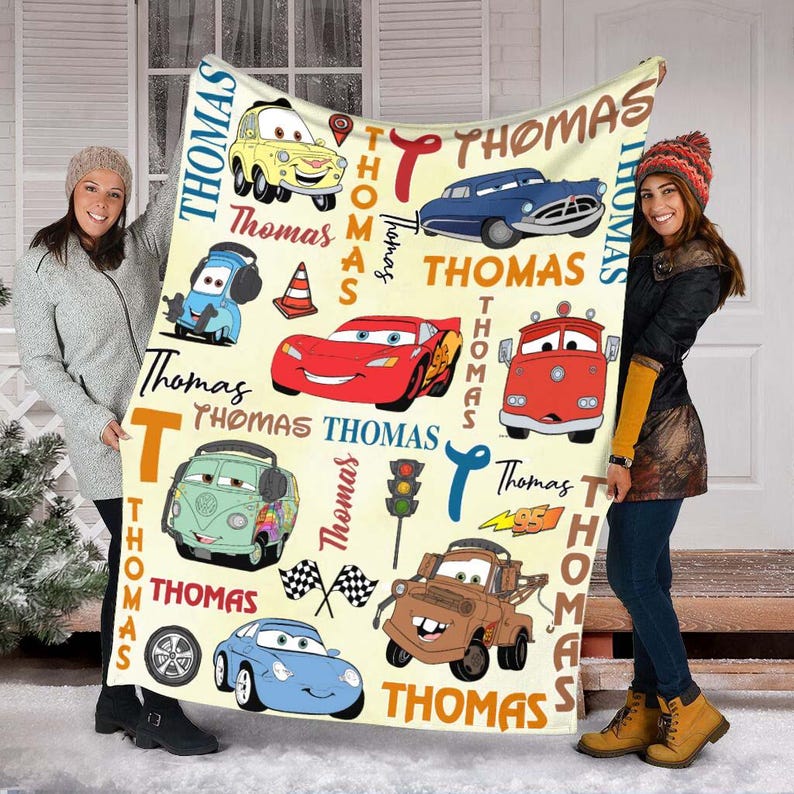 May include: A cream-colored blanket featuring cartoon cars and the name "Thomas" in various fonts and colors. The blanket showcases images of a yellow car, a blue car, a red race car, and a brown tow truck.