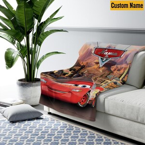 May include: A red Cars-themed blanket featuring Lightning McQueen. The blanket displays the Cars logo and a desert landscape. The words "Custom Name" are in a yellow box in the upper right corner.