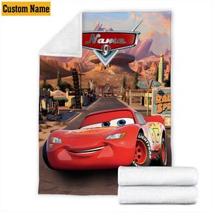 May include: A plush, white blanket featuring a vibrant illustration of Lightning McQueen from the movie Cars. The design includes the character, the Radiator Springs sign, and the text "Name 9". The blanket has a custom name option.