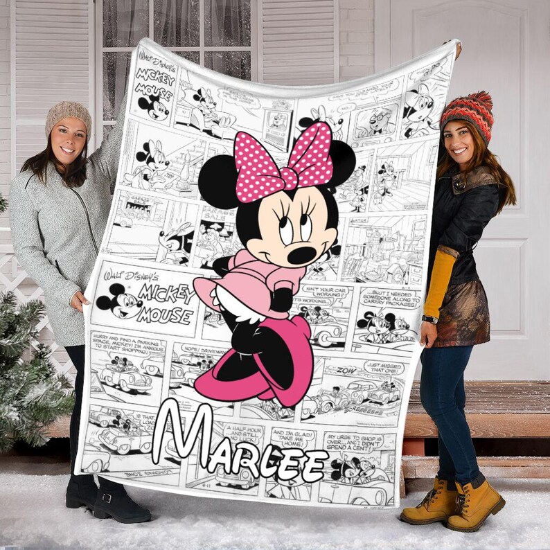 May include: A white fleece blanket featuring a Minnie Mouse comic strip design. The blanket showcases Minnie Mouse in a pink dress and bow, with the name "Marlee" printed below. The blanket is held up by two people.