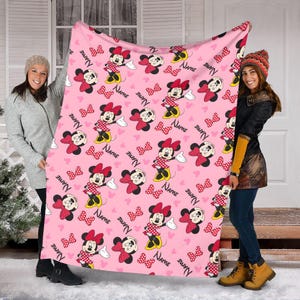 May include: Pink blanket featuring a repeating pattern of Minnie Mouse, bows, and hearts. Minnie Mouse is depicted in her classic red and white polka dot dress and bow. The word "NAME" is also printed on the blanket.