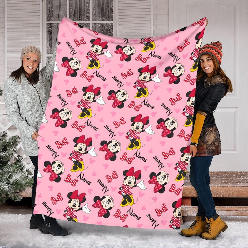 May include: Pink blanket featuring a repeating pattern of Minnie Mouse, bows, and hearts. Minnie Mouse is depicted in her classic red and white polka dot dress and bow. The word "NAME" is also printed on the blanket.