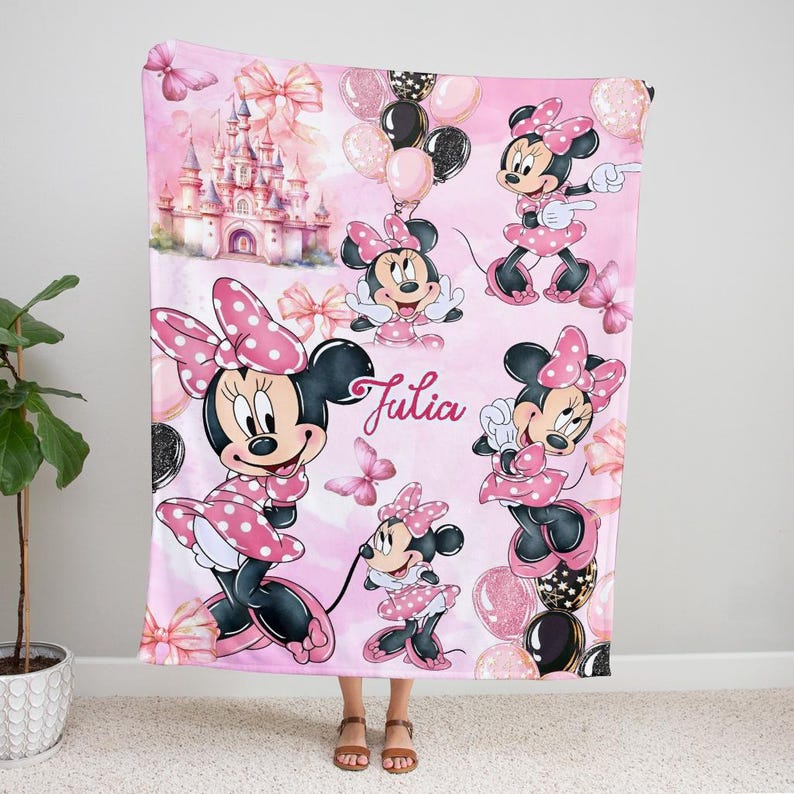 May include: A soft pink fleece blanket featuring multiple illustrations of Minnie Mouse in a polka-dot dress and bow, a large castle, butterflies, and balloons. The name Julia is printed in a cursive script in the centre of the blanket.