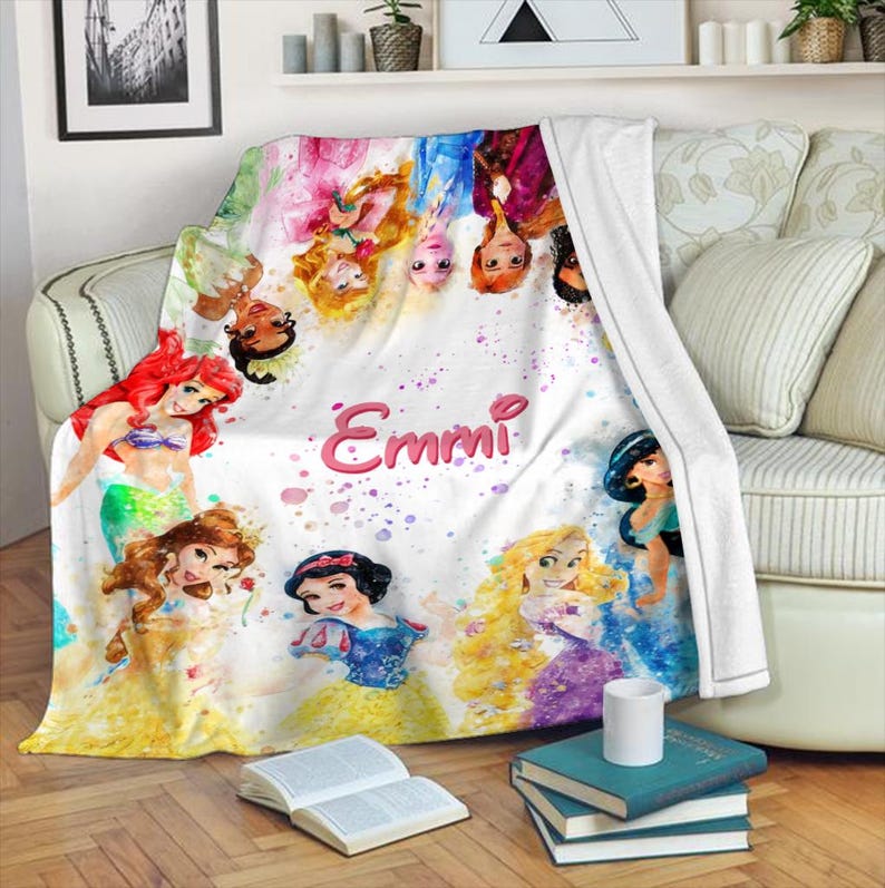May include: A white throw blanket featuring watercolor illustrations of various princesses. The blanket has a personalized name, "Emmi," in pink script. The princesses are surrounded by colorful paint splatters, creating a whimsical design.