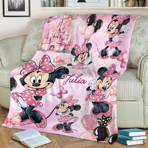 May include: A soft pink fleece blanket featuring a repeating pattern of Minnie Mouse in a polka dot dress, a large castle, pink bows, butterflies, and balloons. The name Julia is printed in a cursive script in the centre of the blanket.