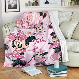 May include: A soft pink fleece blanket featuring a repeating pattern of Minnie Mouse in a polka dot dress, a large castle, balloons, and butterflies. The name Julia is printed in a cursive script in the centre of the blanket design.