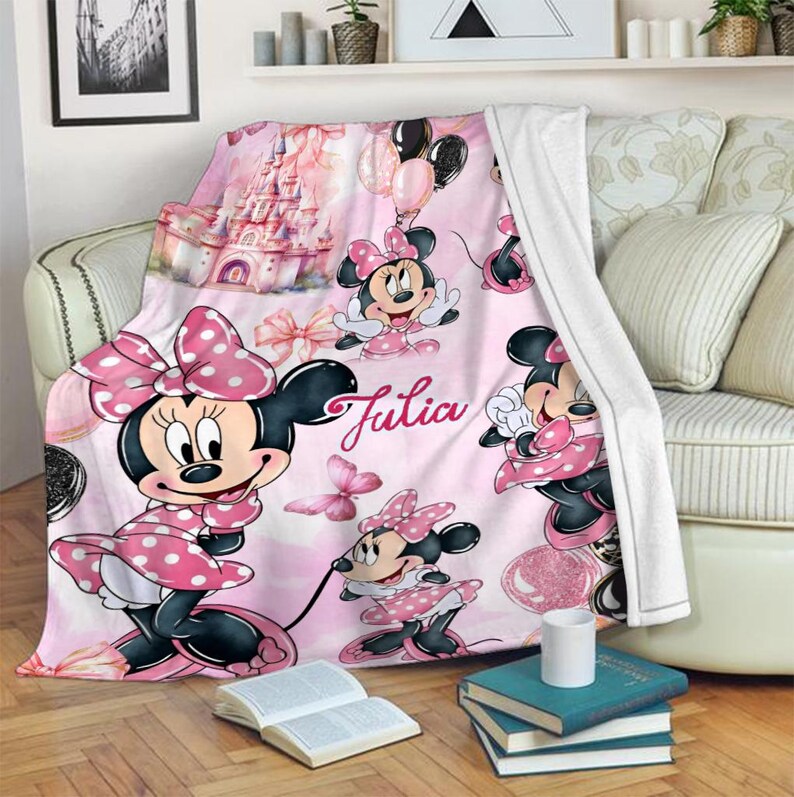 May include: A soft pink fleece blanket featuring a repeating pattern of Minnie Mouse in a polka dot dress, a large castle, balloons, and butterflies. The name Julia is printed in a cursive script in the centre of the blanket design.