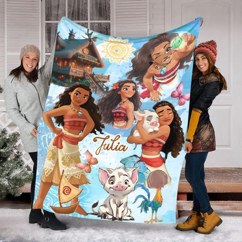 May include: A soft fleece blanket featuring a collage of Moana characters, including Moana, Pua the pig, and Heihei the rooster, alongside a tropical hut, a sailboat, and a sun. The name Julia is printed in brown script in the center of the design.