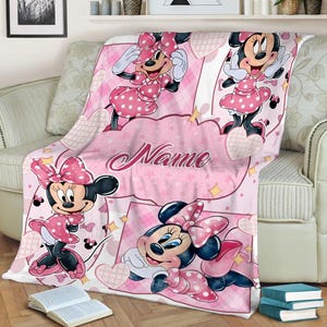 May include: A soft pink fleece blanket featuring multiple illustrations of Minnie Mouse wearing a polka dot dress and bow. The design includes heart shapes, stars, and a central area with the word Name written in a cursive script font.