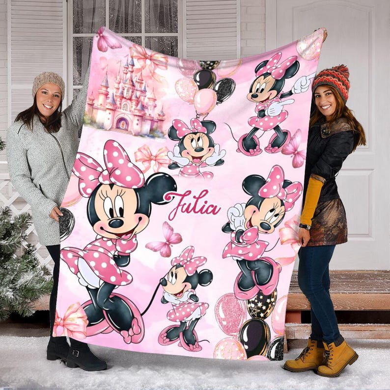 May include: A soft pink fleece blanket featuring multiple illustrations of Minnie Mouse in a polka-dot dress, a fairy tale castle, balloons, and butterflies. The name Julia is printed in a cursive script in the centre of the blanket.