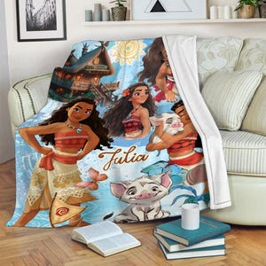 May include: A soft fleece blanket featuring a blue background with illustrations of the character Moana, a small pig, and a chicken. The design includes a tropical hut, a butterfly, and the name Julia written in a brown cursive script font.