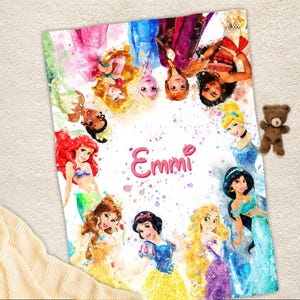 May include: A personalized blanket featuring watercolor illustrations of various princesses. The blanket has a white background with the name "Emmi" in pink script. A small brown teddy bear is placed on the side of the blanket.