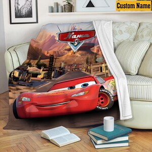 May include: A red Lightning McQueen blanket with a desert landscape background. The blanket features the text "Name" and the number 9. The blanket is draped over a sofa with white pillows and a white throw.