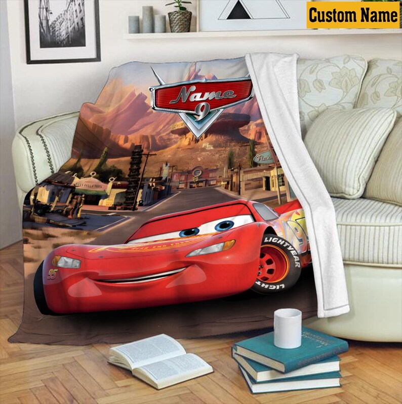 May include: A red Lightning McQueen blanket with a desert landscape background. The blanket features the text "Name" and the number 9. The blanket is draped over a sofa with white pillows and a white throw.