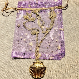 May include: A gold-colored seashell pendant necklace with a delicate chain, resting on a small purple drawstring bag. The bag has a celestial design with moons and stars. The necklace is a jewelry item.