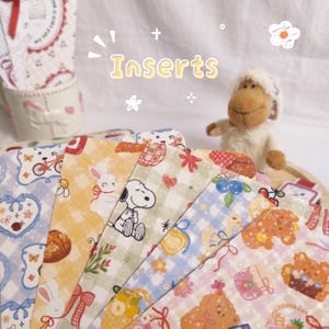 Op de afbeelding: Een verzameling kleurrijke inlegvellen met verschillende patronen, waaronder een fiets, konijn, Snoopy en teddybeer. De inlegvellen zijn in een waaier gerangschikt, met het woord "Inserts" in het geel. Een pluchen schaapje staat op de achtergrond.