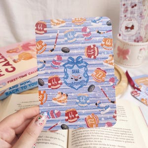 Peut inclure: Une carte rectangulaire avec un fond bleu clair présentant un motif de maillots de hockey, de patins et de palets. La carte porte le texte "Briar University" dans un motif de blason. La carte est tenue au-dessus d'un livre ouvert.