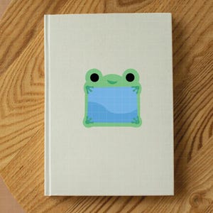 May include: A white hardcover notebook featuring a centered graphic of a light green frog with large black eyes. The frog is holding a square blue frame with a grid pattern and a wavy line across the bottom, creating a simple, clean illustration.