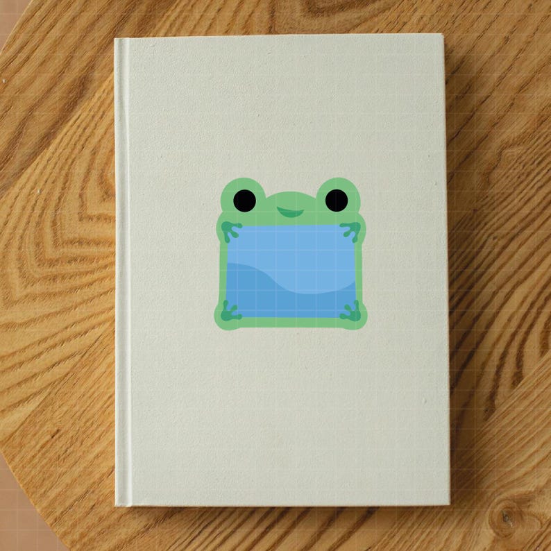May include: A white hardcover notebook featuring a centered graphic of a light green frog with large black eyes. The frog is holding a square blue frame with a grid pattern and a wavy line across the bottom, creating a simple, clean illustration.