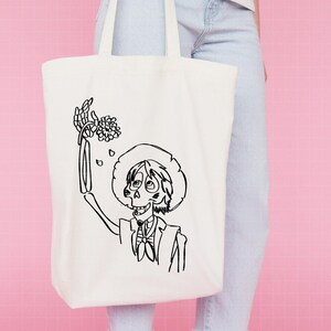 Puede incluir: Bolso tote de color crema con una ilustraci&oacute;n en blanco y negro de un personaje esqueleto con sombrero y traje, sosteniendo flores. El personaje est&aacute; sonriendo. El bolso tiene dos asas y est&aacute; hecho de un material similar a la lona.