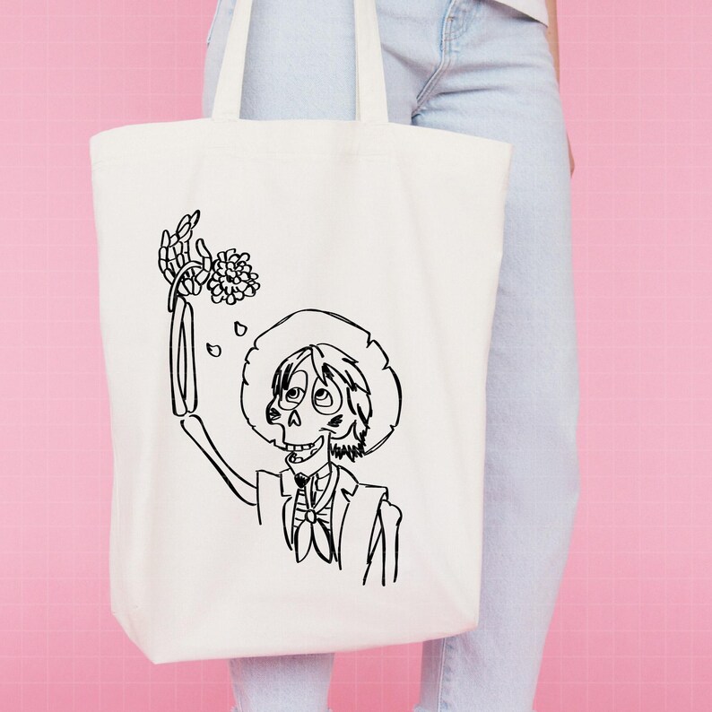 Puede incluir: Bolso tote de color crema con una ilustraci&oacute;n en blanco y negro de un personaje esqueleto con sombrero y traje, sosteniendo flores. El personaje est&aacute; sonriendo. El bolso tiene dos asas y est&aacute; hecho de un material similar a la lona.