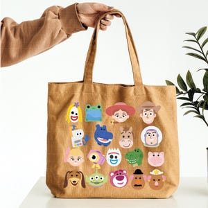 May include: A tan canvas tote bag featuring a grid of colorful cartoon character faces and icons. The design includes various animated figures such as a cowboy, a space ranger, a dinosaur, a pig, a potato character, and an alien, all printed in a flat style.