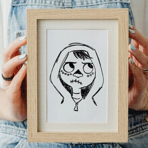 Puede incluir: Un boceto en blanco y negro enmarcado de un personaje con rostro de calavera, que lleva una sudadera con capucha. La obra de arte est&aacute; en un marco de madera de color claro. El personaje tiene ojos grandes y una boca cosida.