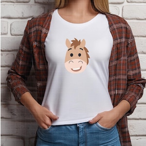 May include: A white short-sleeved t-shirt featuring a centered graphic of a smiling brown horse head with a dark brown mane, worn under an unbuttoned long-sleeved brown and red plaid flannel shirt paired with blue denim jeans.