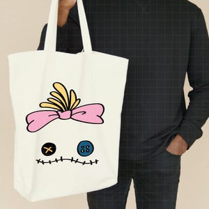Pu&ograve; includere: Borsa tote color crema con un design a volto stravagante. Il volto include un fiocco rosa, accenti gialli, un occhio nero a forma di croce, un occhio a bottone blu e una bocca cucita. La borsa &egrave; tenuta da una persona.