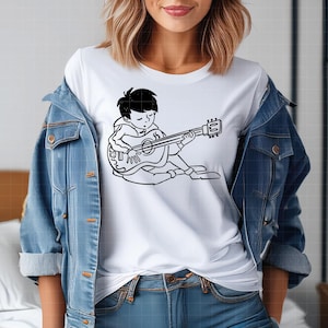 Puede incluir: Camiseta blanca con una ilustraci&oacute;n en blanco y negro de una persona tocando una guitarra ac&uacute;stica. El gr&aacute;fico muestra una figura sentada con los ojos cerrados, sosteniendo la guitarra. La camiseta se usa con una chaqueta vaquera azul.