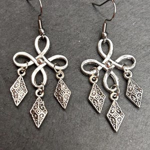 May include: A pair of silver-toned dangle earrings. Each earring features a decorative cross-like design with three dangling diamond-shaped charms. The charms have an ornate, patterned surface. The earrings have a hook closure.
