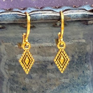 May include: A pair of gold-colored earrings with a diamond-shaped pendant. The earrings are hanging from a blue ceramic vase. The diamond pendants have a textured design and a small yellow center.