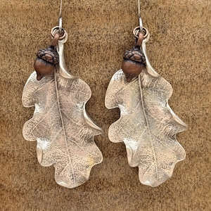 May include: A pair of dangle earrings featuring textured silver-toned oak leaf charms. Each leaf is accented with a small, copper-toned acorn charm attached to a silver-toned ear wire. The metal surfaces show detailed veining and a matte finish.