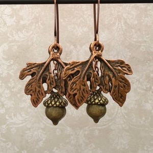 May include: A pair of dangle earrings featuring copper-toned metal oak leaves with textured surfaces. Each earring includes a small, brass-colored acorn charm suspended from the center of the leaf design, attached to a simple copper-toned metal hook.