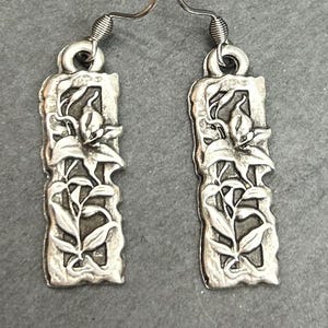May include: A pair of silver-toned dangle earrings. Each earring features a rectangular design with a floral and leaf motif. The earrings have a hook closure and are displayed against a gray background.