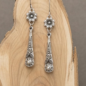 May include: Silver-toned dangle earrings. Each earring features a floral design at the top, a long, ornate pendant with intricate patterns, and a small, round bead at the bottom. The earrings are displayed on a wooden surface.