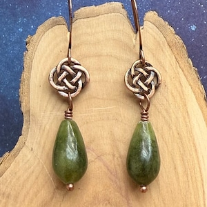 May include: A pair of copper-colored Celtic knot earrings with teardrop-shaped green jade stones. The earrings have a hook closure and are displayed on a wooden surface. The earrings are a unique piece of jewelry.