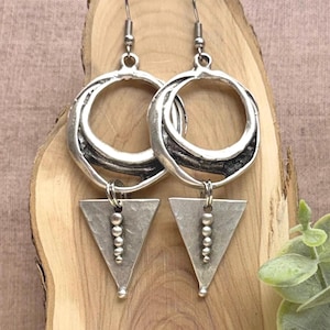 May include: A pair of silver-toned dangle earrings featuring a circular hoop top with a textured, dark recessed center. Suspended from each hoop is a flat, downward-pointing triangle pendant adorned with a vertical row of five small silver beads.