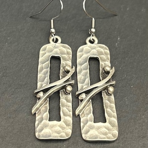 May include: A pair of silver-toned earrings. Each earring has a rectangular shape with a hammered texture and a central cutout. Two diagonal bars and three small spheres adorn each earring. The earrings have a hook closure.