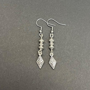 May include: A pair of silver dangle earrings. Each earring features a decorative diamond-shaped charm with an ornate pattern, suspended below a series of beads and a hook. The earrings are displayed against a dark background.