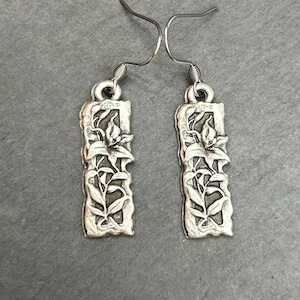 May include: A pair of silver-toned dangle earrings. Each earring features a rectangular frame with a floral design. The earrings have a textured, antique appearance and are suspended from curved ear wires.