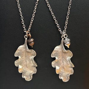 May include: Two silver-toned leaf pendant necklaces with acorn charms. The pendants are detailed with leaf veins and hang from delicate chain necklaces. The acorns are a bronze color. The background is black.