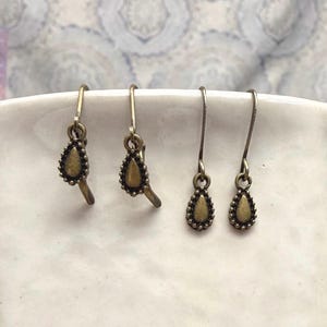 May include: Four pairs of antique brass-colored earrings. Each earring features a teardrop-shaped pendant with a beaded border and a small, curved extension. The earrings are displayed on a white surface.
