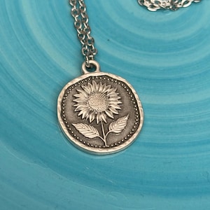 May include: A silver-toned sunflower pendant necklace. The pendant features a detailed sunflower design with leaves and a dotted border. The necklace chain is also silver and visible against a blue background.