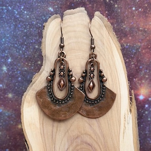 May include: A pair of copper-colored earrings with a fan-shaped design. The earrings feature intricate detailing, including small beads and a central diamond-shaped element. The earrings are displayed on a wooden surface against a starry background.