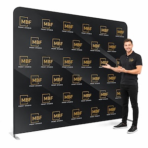May include: A large black backdrop with a repeating gold logo that reads "MBF PRINT STUDIO." The backdrop is supported by a silver metal frame. A person in a black polo shirt and black pants stands next to the backdrop, gesturing towards it.